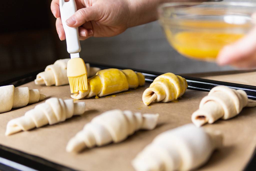 Baking croissants at home and covering the dough with egg yolk using pastry brush |
